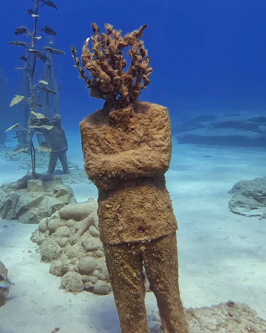 An underwater statue of a man with coral growing from his head, arms crossed